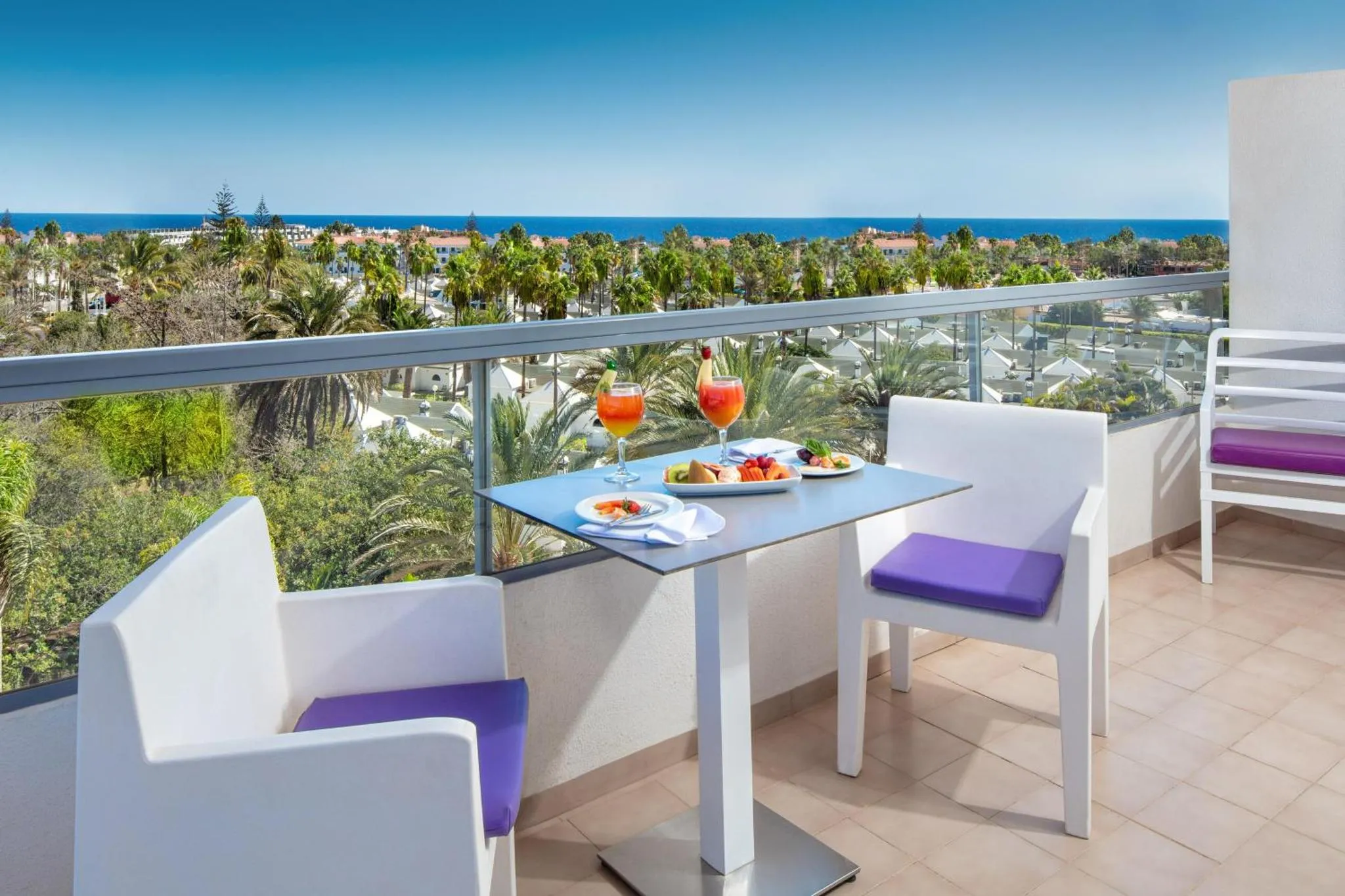 Une table avec des boissons et des fruits est mise en scène sur une terrasse avec vue sur la mer et la végétation.