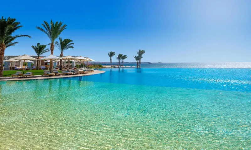 Une piscine à débordement turquoise bordée de transats sous des parasols, avec une mer bleue en arrière-plan.