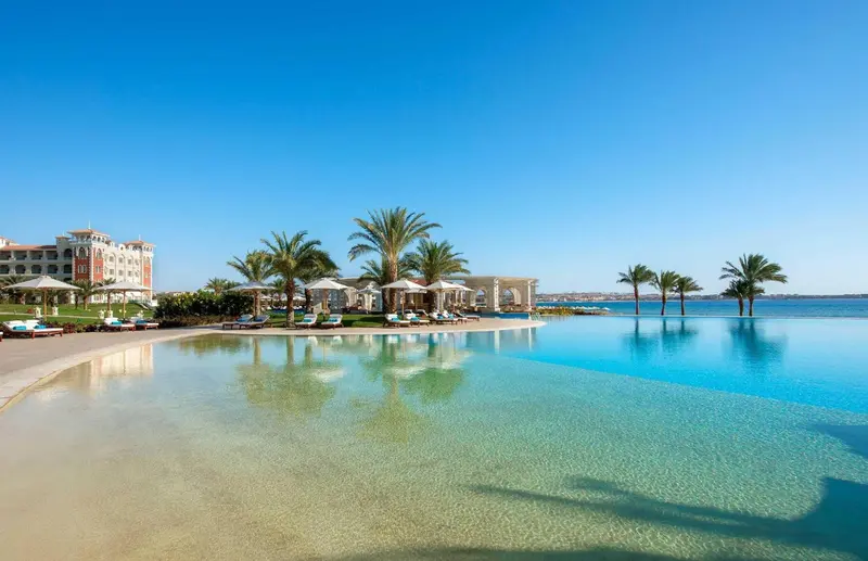 Une piscine à débordement turquoise s'étend devant un bâtiment blanc et des palmiers.