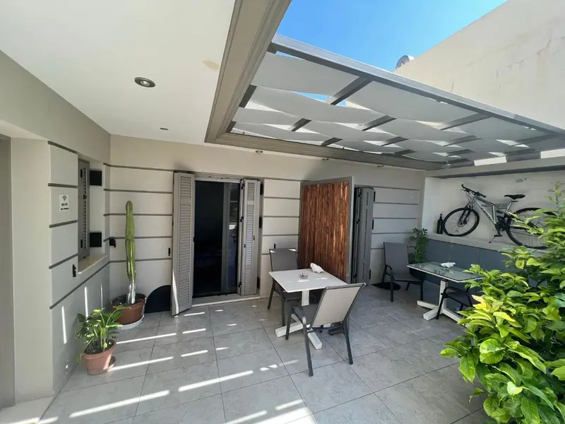 Terrasse ombragée avec table et chaises, devant une porte ouverte.