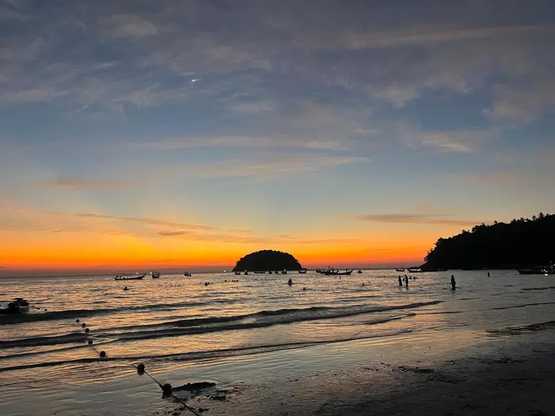 Coucher de soleil sur une plage avec des bateaux et des silhouettes de personnes.