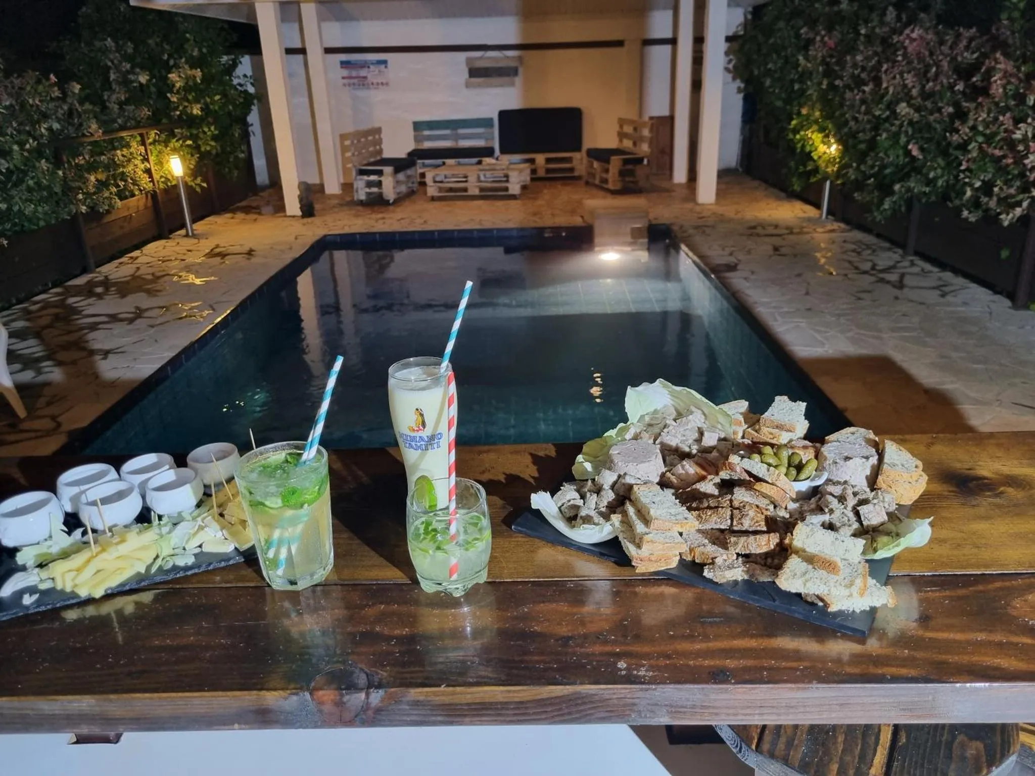Une table est dressée au bord d'une piscine illuminée, avec des boissons et des amuse-gueules.