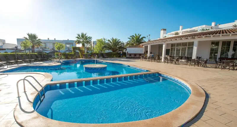 Piscine bleue entourée d'un sol carrelé et de chaises longues, avec des bâtiments blancs en arrière-plan.