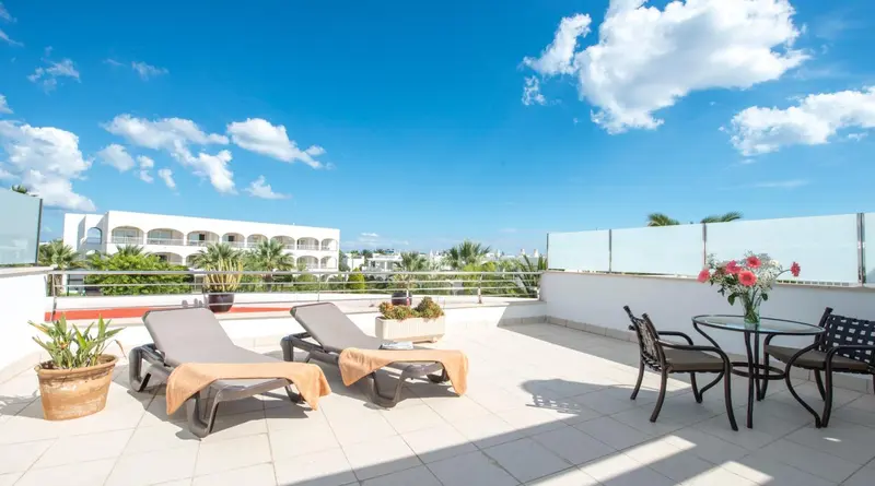 Terrasse aménagée avec transats et table basse, vue sur un bâtiment blanc et un ciel bleu nuageux.
