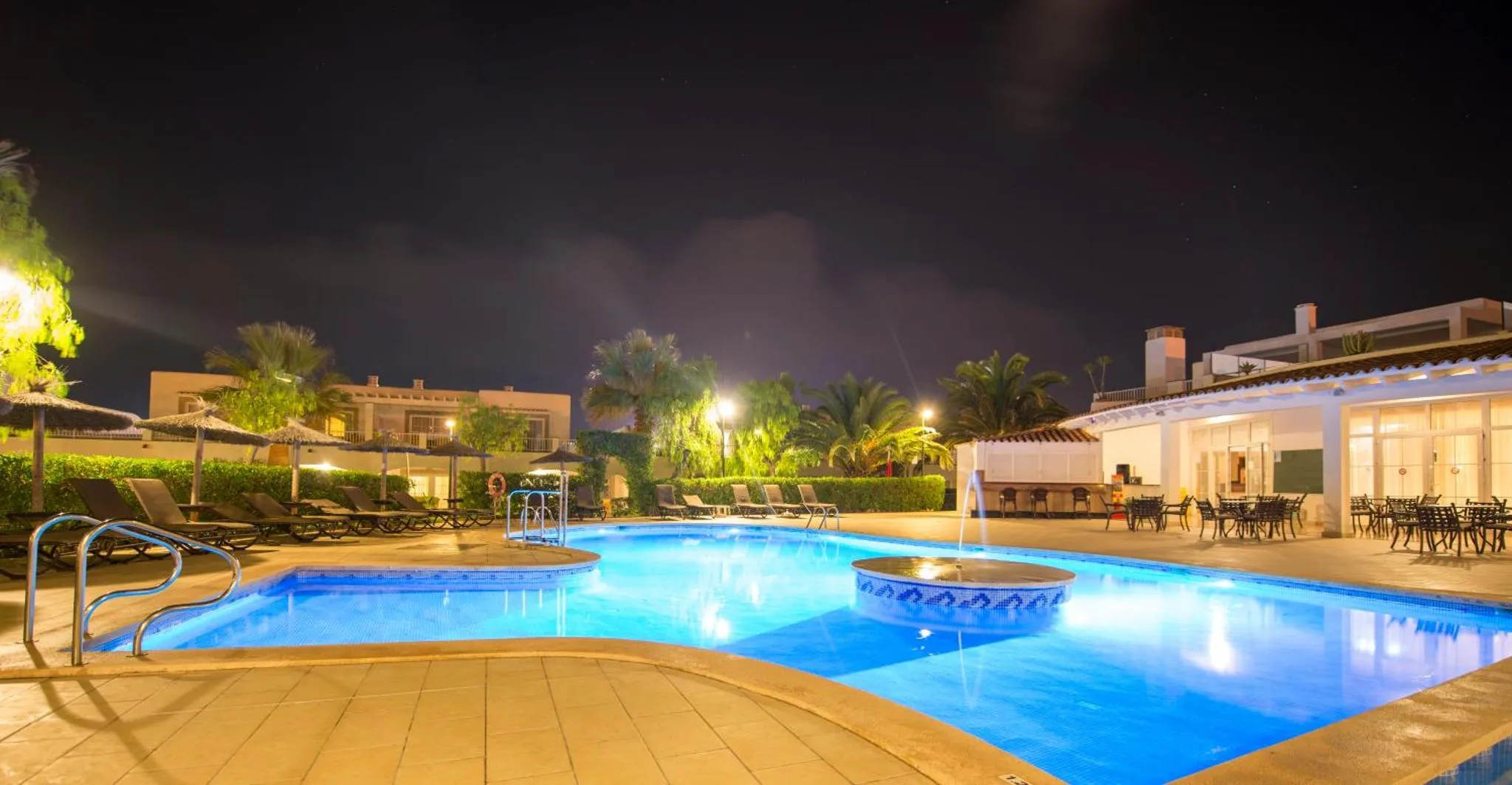 Piscine illuminée avec transats et végétation tropicale la nuit.