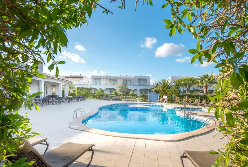 Une piscine rectangulaire bordée de chaises longues et de végétation luxuriante, devant un bâtiment blanc.