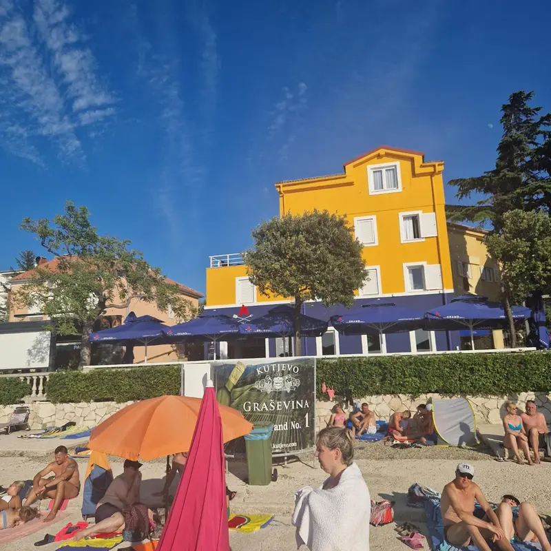 Baigneurs sur une plage ensoleillée devant un bâtiment jaune et des parasols.