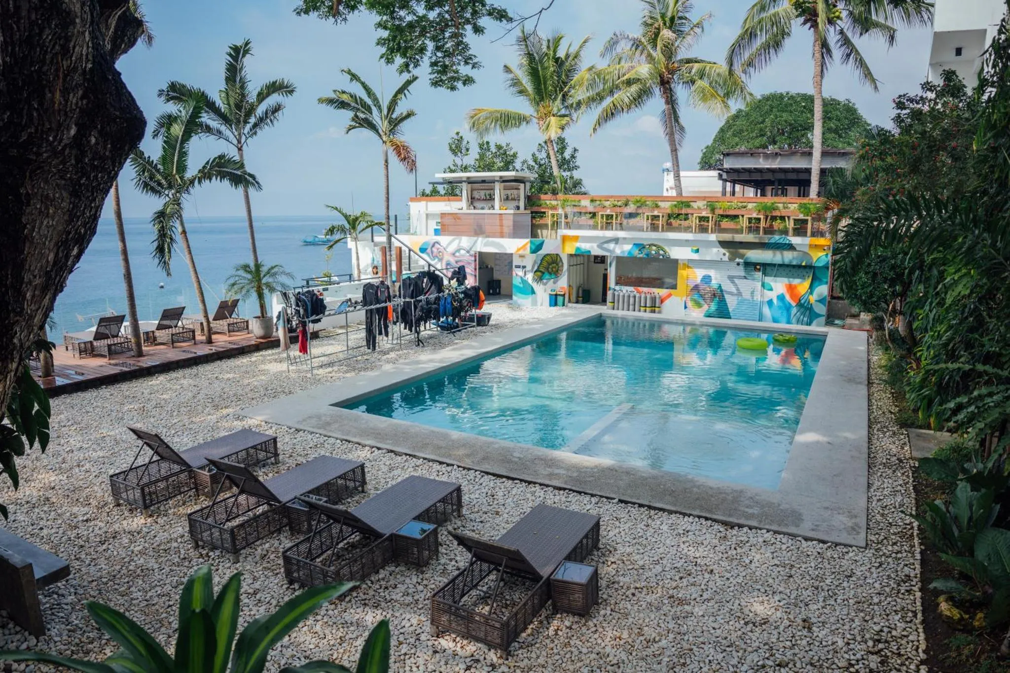 Une piscine rectangulaire bordée de chaises longues, avec un bâtiment coloré en arrière-plan et la mer au loin.