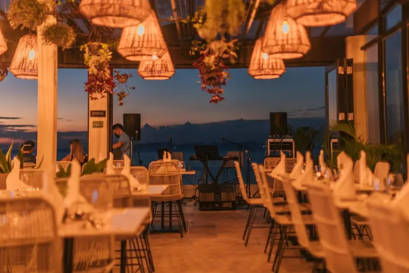 Une terrasse lumineuse avec des tables dressées face à un paysage marin au crépuscule.