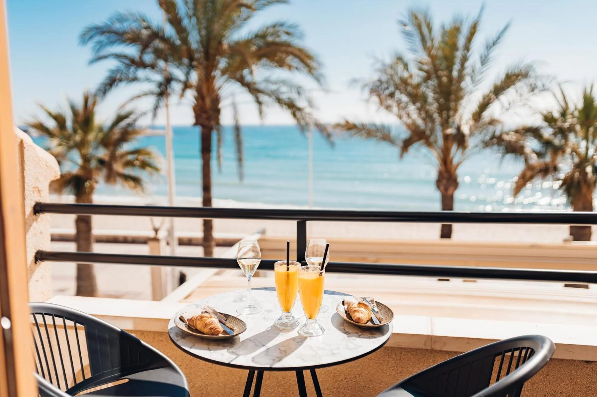 Une table ronde avec des boissons et des viennoiseries sur une terrasse ensoleillée avec vue sur la mer et des palmiers.