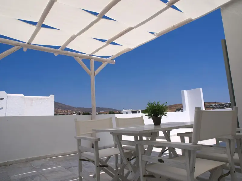 Une table et des chaises blanches sont disposées sur une terrasse ombragée avec vue sur un paysage montagneux.