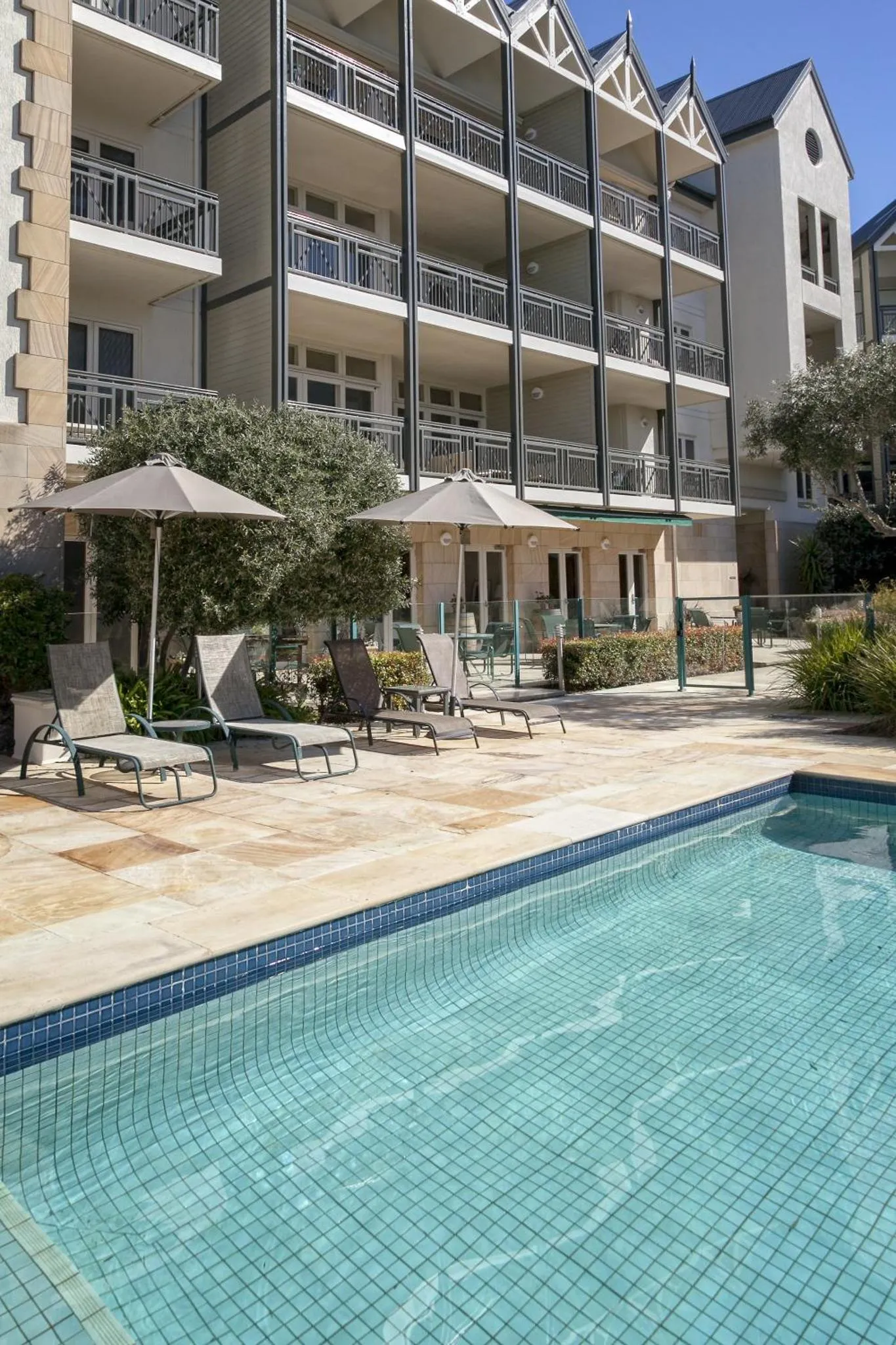Piscine turquoise bordée d'une terrasse avec transats et parasols devant un bâtiment aux balcons.