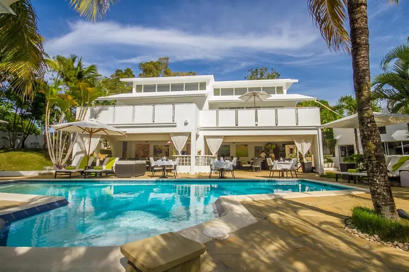 Une piscine turquoise bordée de chaises longues devant un bâtiment blanc.