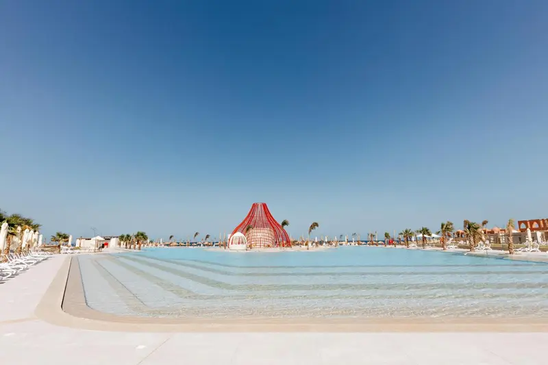 Une plage de sable fin borde une piscine turquoise sous un ciel bleu dégagé.