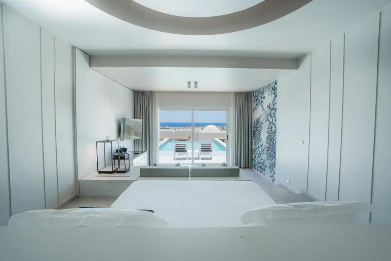 Une chambre lumineuse avec un grand lit blanc et une vue sur une piscine et la mer.