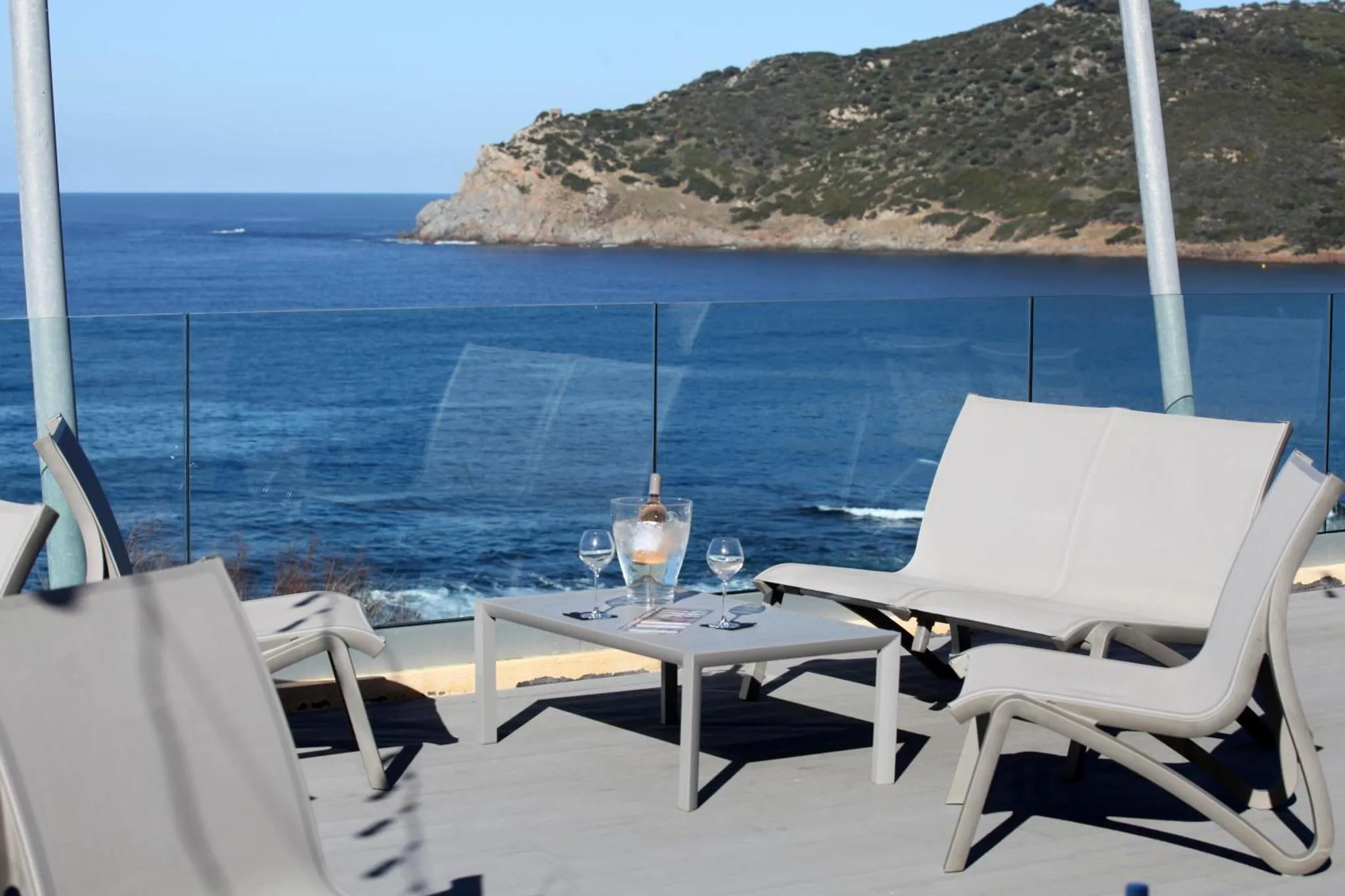 Transats et table basse avec boissons sur une terrasse ensoleillée face à la mer.
