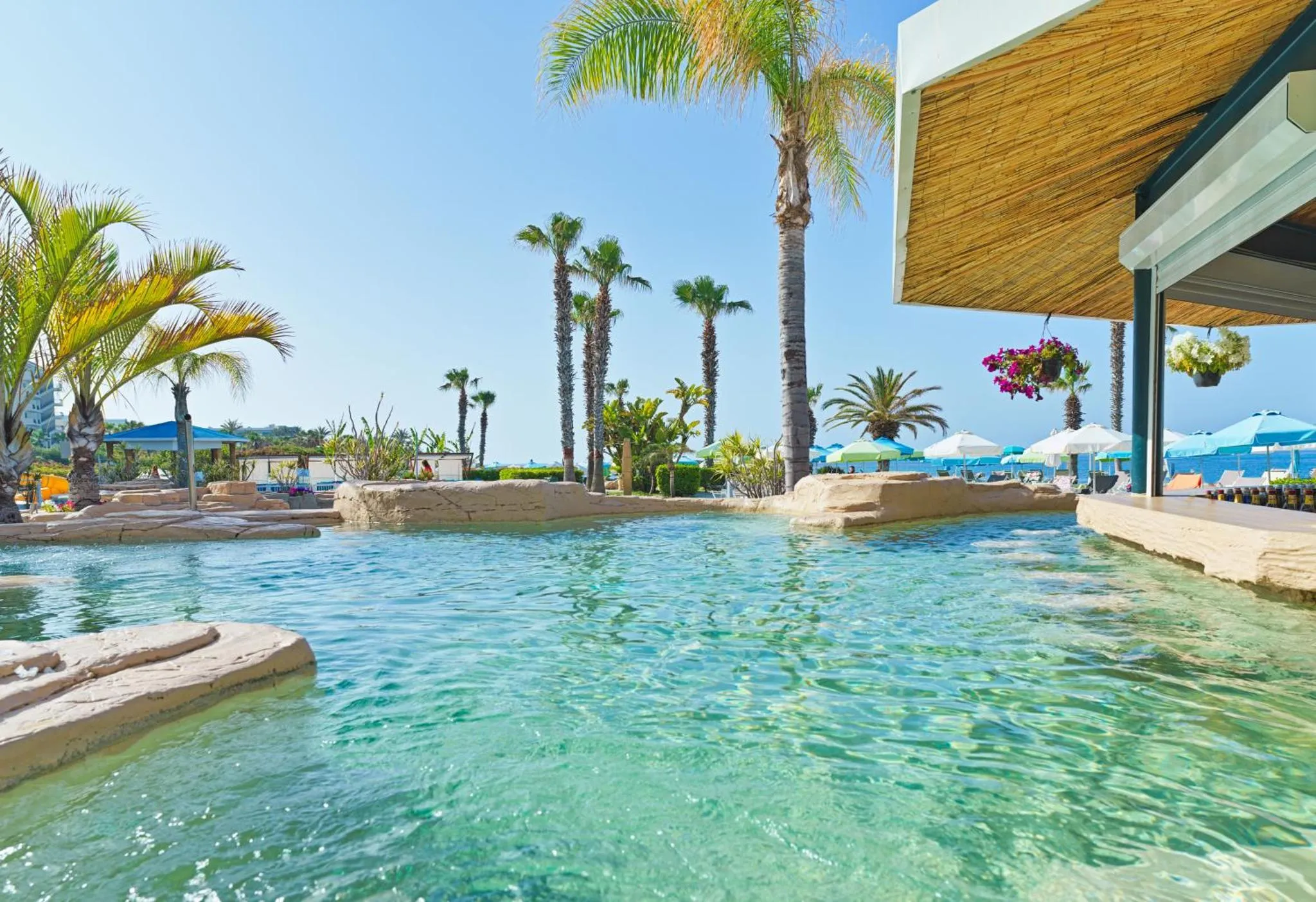 Une piscine turquoise bordée de rochers et de végétation tropicale, avec des transats et parasols en arrière-plan.