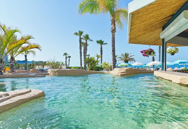Une piscine turquoise bordée de rochers et de végétation tropicale, avec des transats et parasols en arrière-plan.