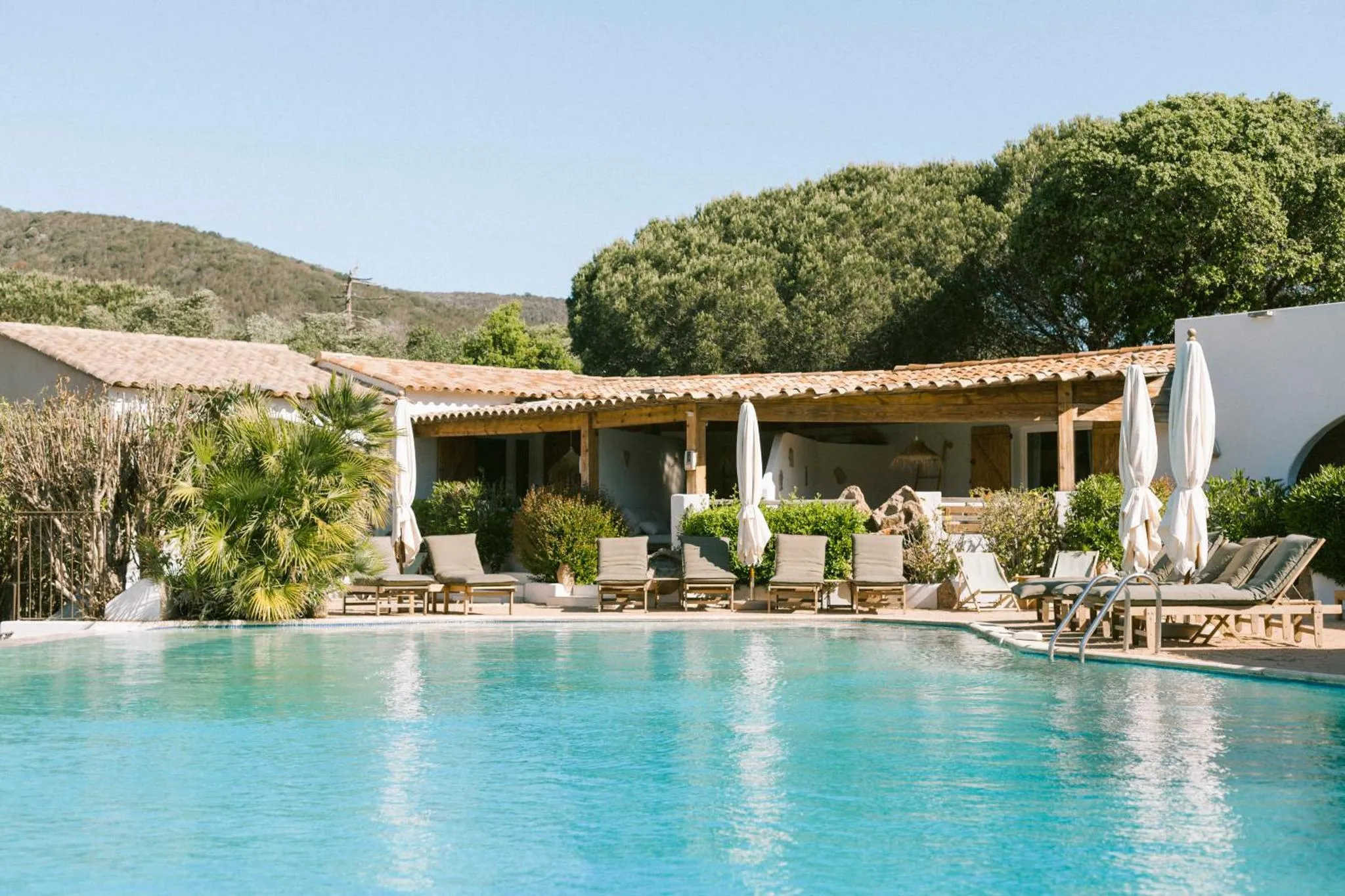 Une piscine turquoise bordée de transats et de parasols, devant un bâtiment à colonnes et une végétation luxuriante.