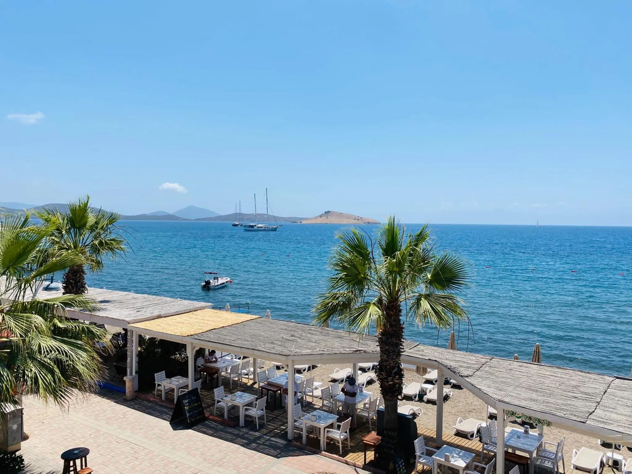 Terrasse ombragée avec tables et chaises donnant sur la mer et des îles au loin.