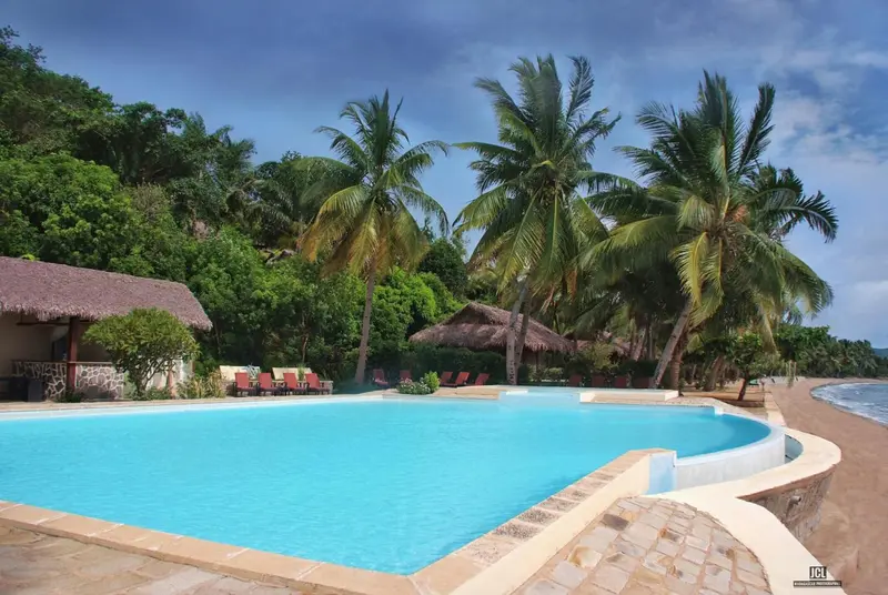 Piscine turquoise bordée d'une terrasse ensoleillée avec des palmiers en arrière-plan.