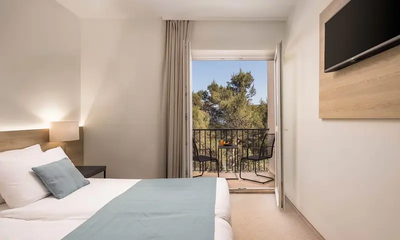 Chambre lumineuse avec un lit fait et un balcon donnant sur la végétation.