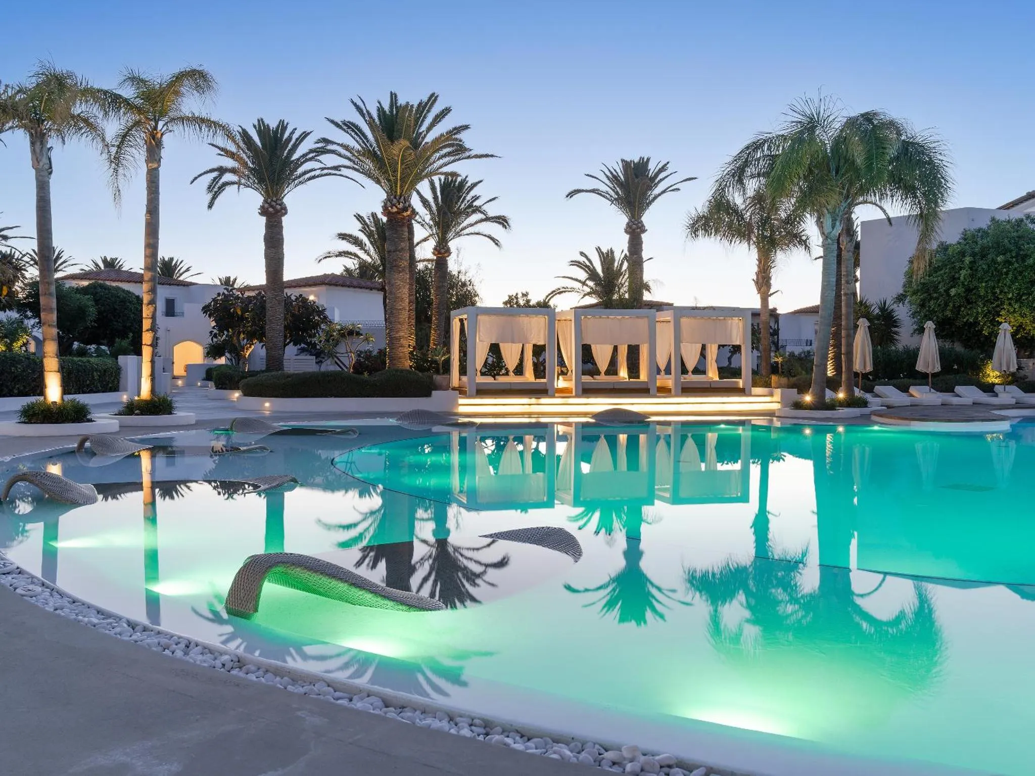 Piscine illuminée avec des palmiers et des transats blancs au crépuscule.