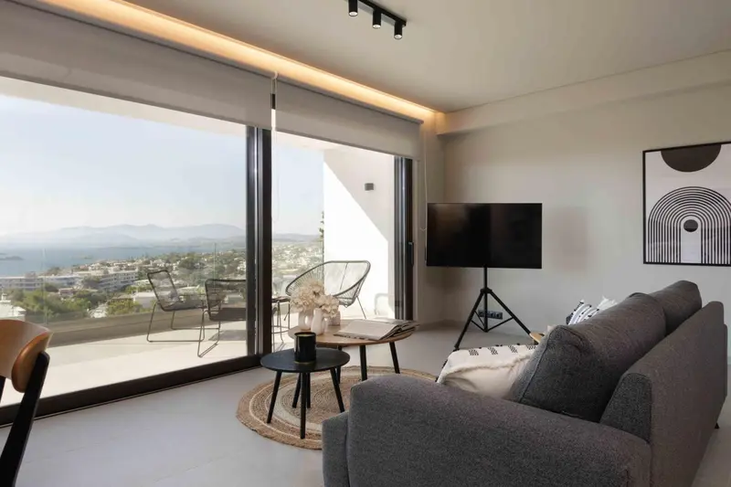 Un salon moderne avec un canapé gris et une grande fenêtre offrant une vue sur la mer et les montagnes.