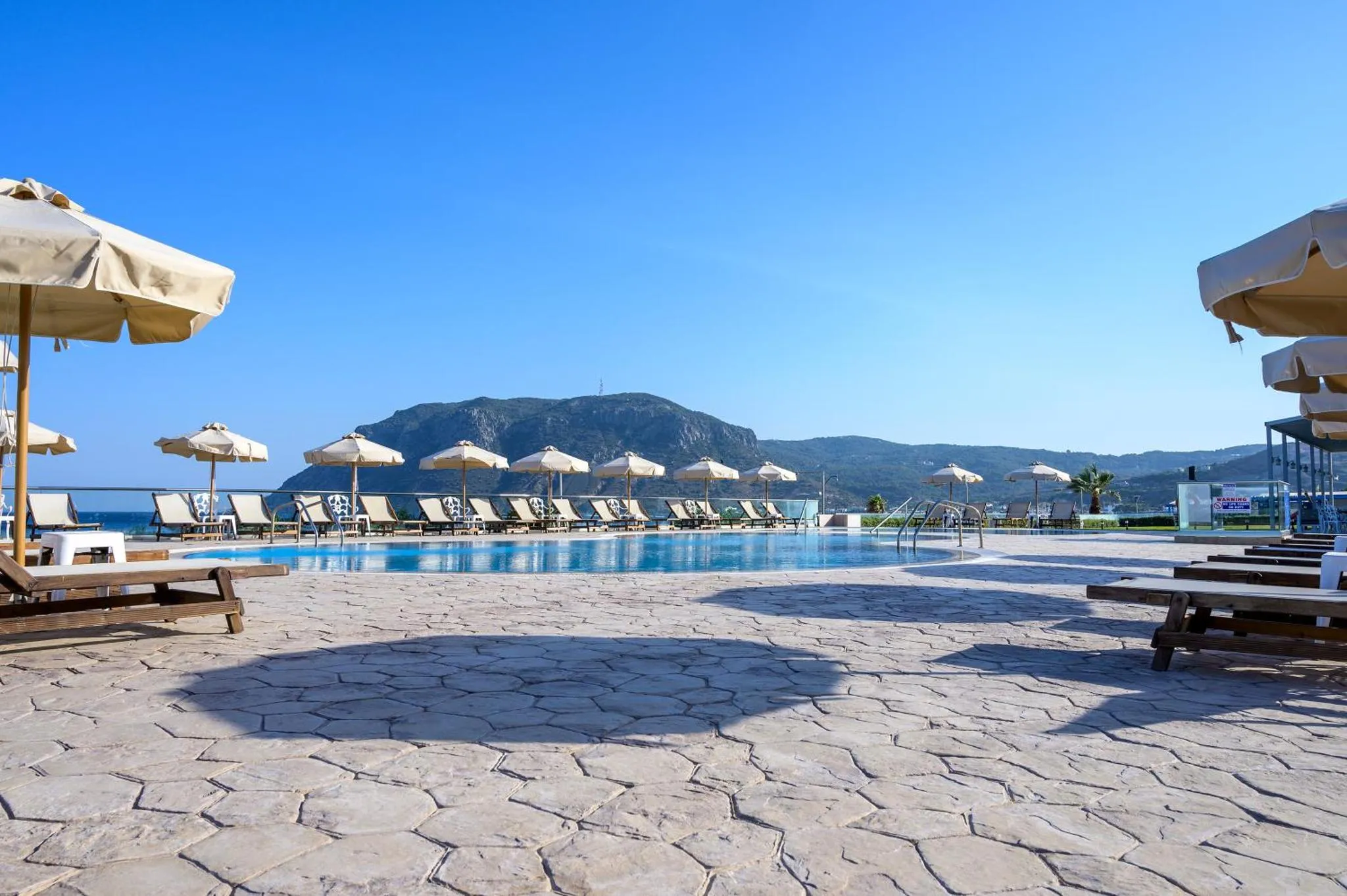 Transats et parasols bordent une piscine ensoleillée avec vue sur la mer et les montagnes.