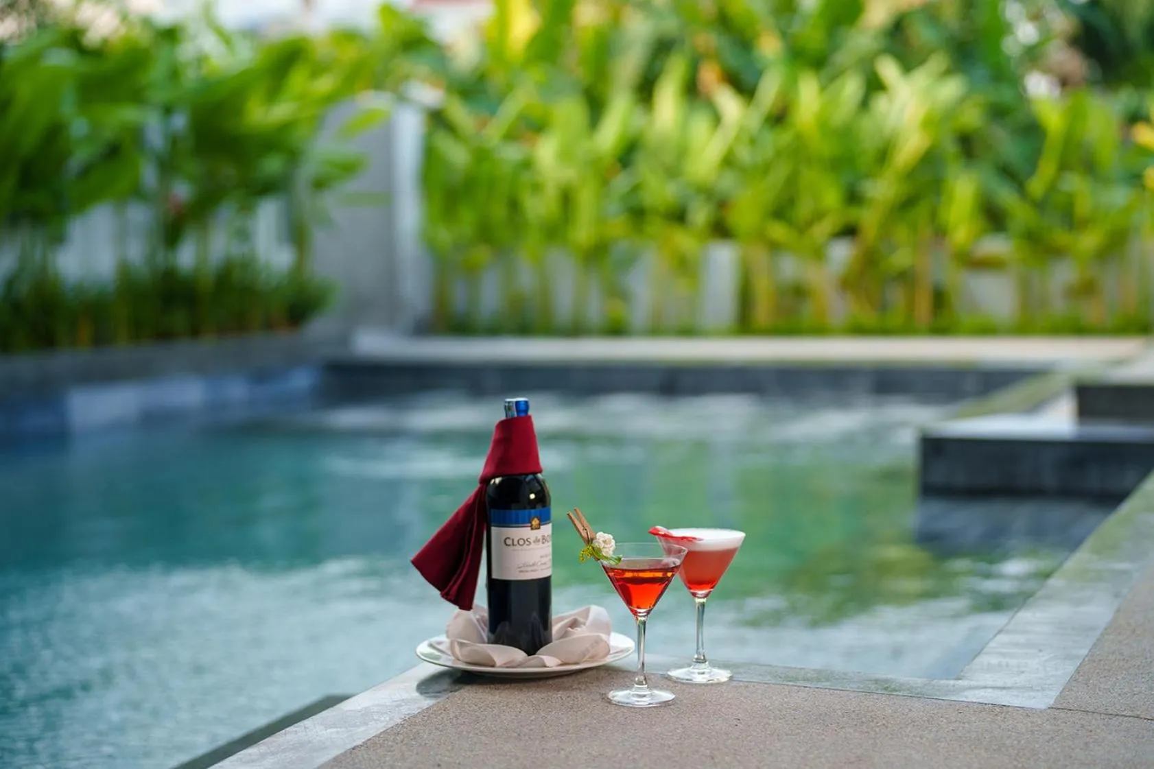Une bouteille de vin et deux verres colorés sont posés sur le bord d'une piscine.