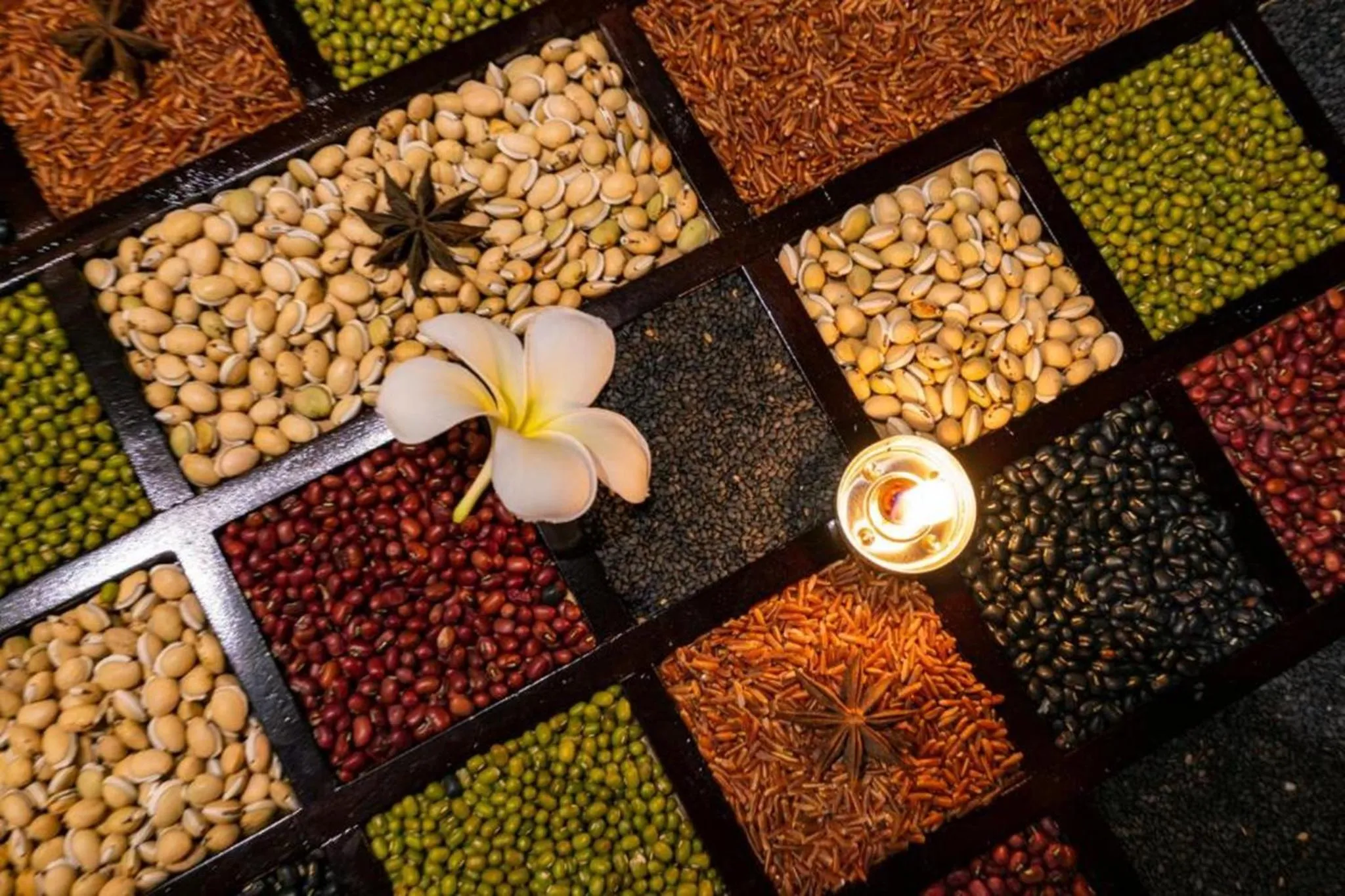 Assortiment de graines et de légumineuses dans des compartiments en bois, décoré d'une fleur et d'une bougie.