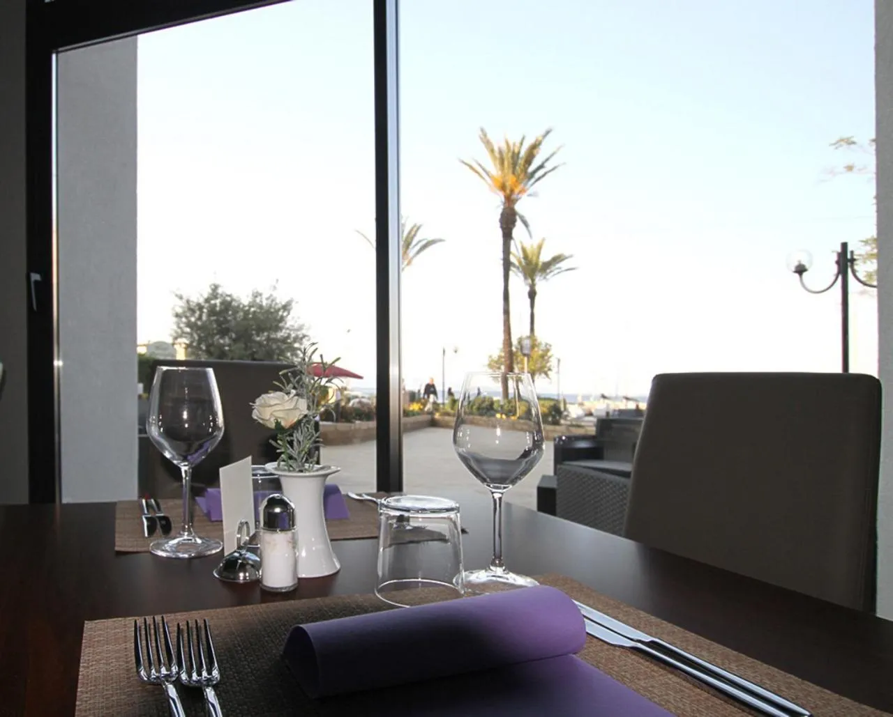 Une table dressée avec couverts et verres face à une vue sur la mer et la végétation.