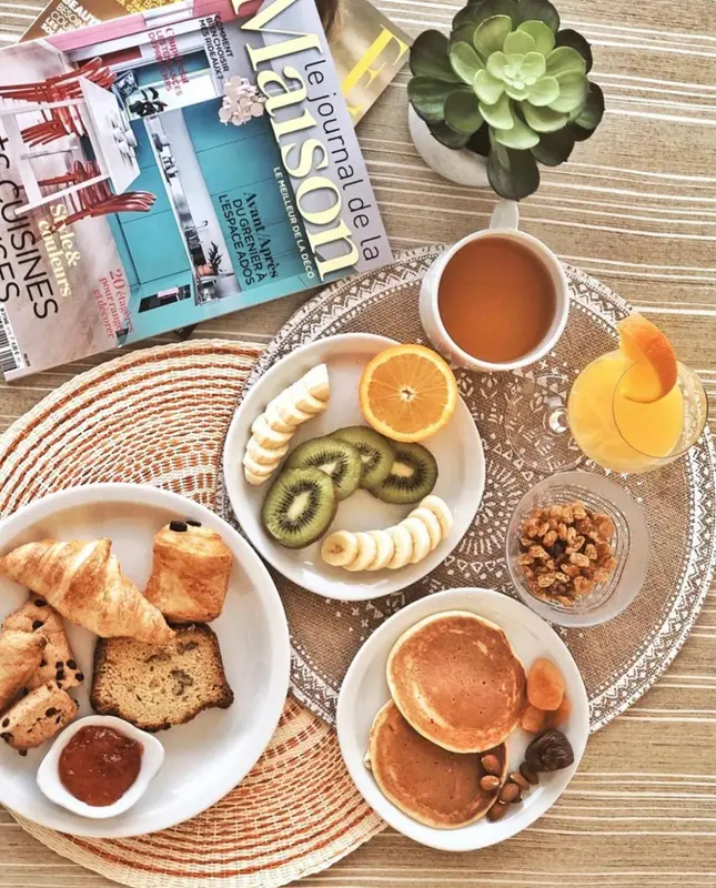 Une table en osier présente un petit-déjeuner avec des fruits frais, des viennoiseries et des crêpes.
