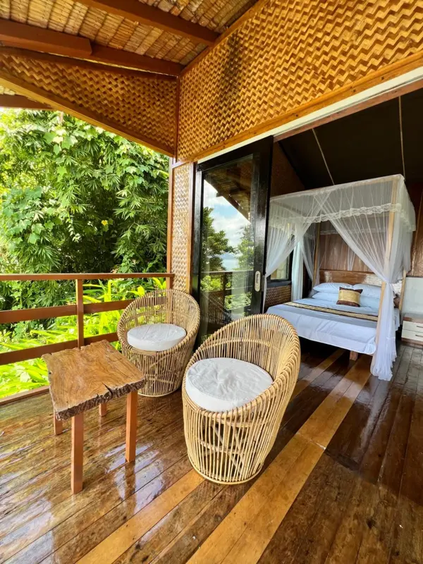 Une terrasse en bois abrite un espace de repos avec des fauteuils en rotin et un lit à moustiquaire.
