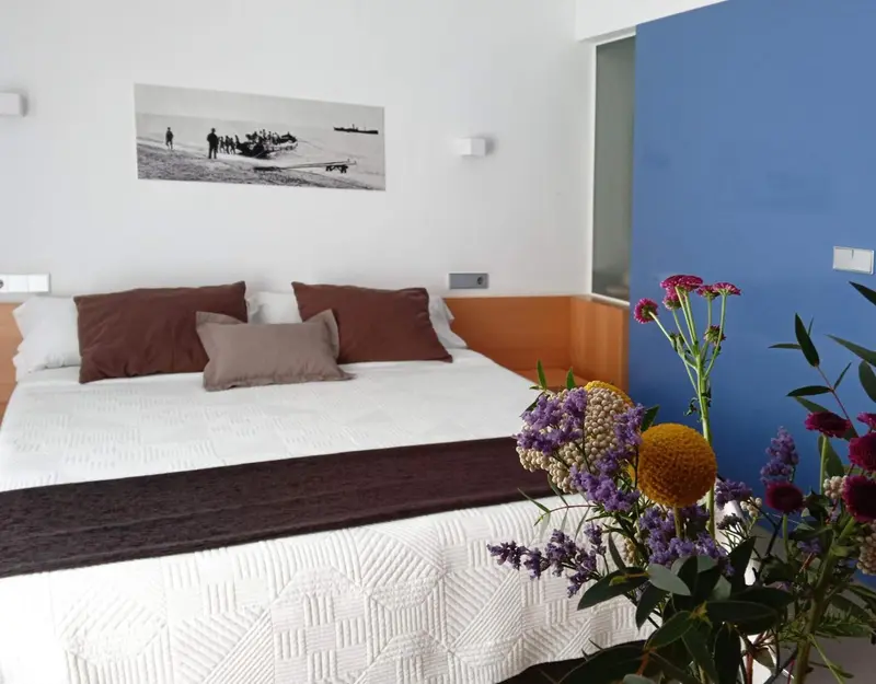 Une chambre lumineuse avec un grand lit décoré de coussins et un bouquet de fleurs.