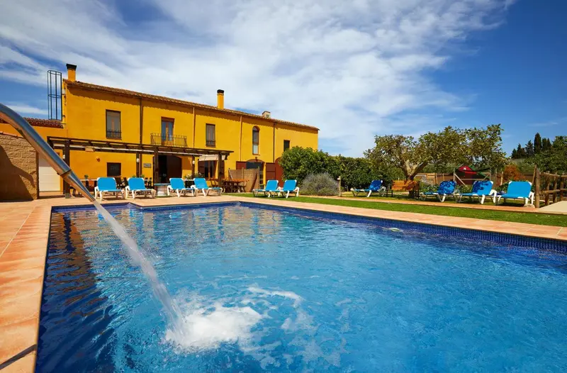 Une piscine turquoise au premier plan, avec un bâtiment jaune et des transats bleus en arrière-plan.