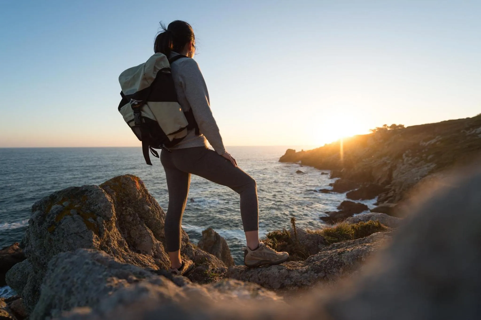 Une personne se tient au bord d'une falaise rocheuse face à la mer au coucher du soleil.