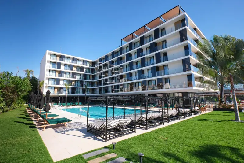 Bâtiment moderne blanc avec des balcons donnant sur une piscine et un jardin verdoyant.