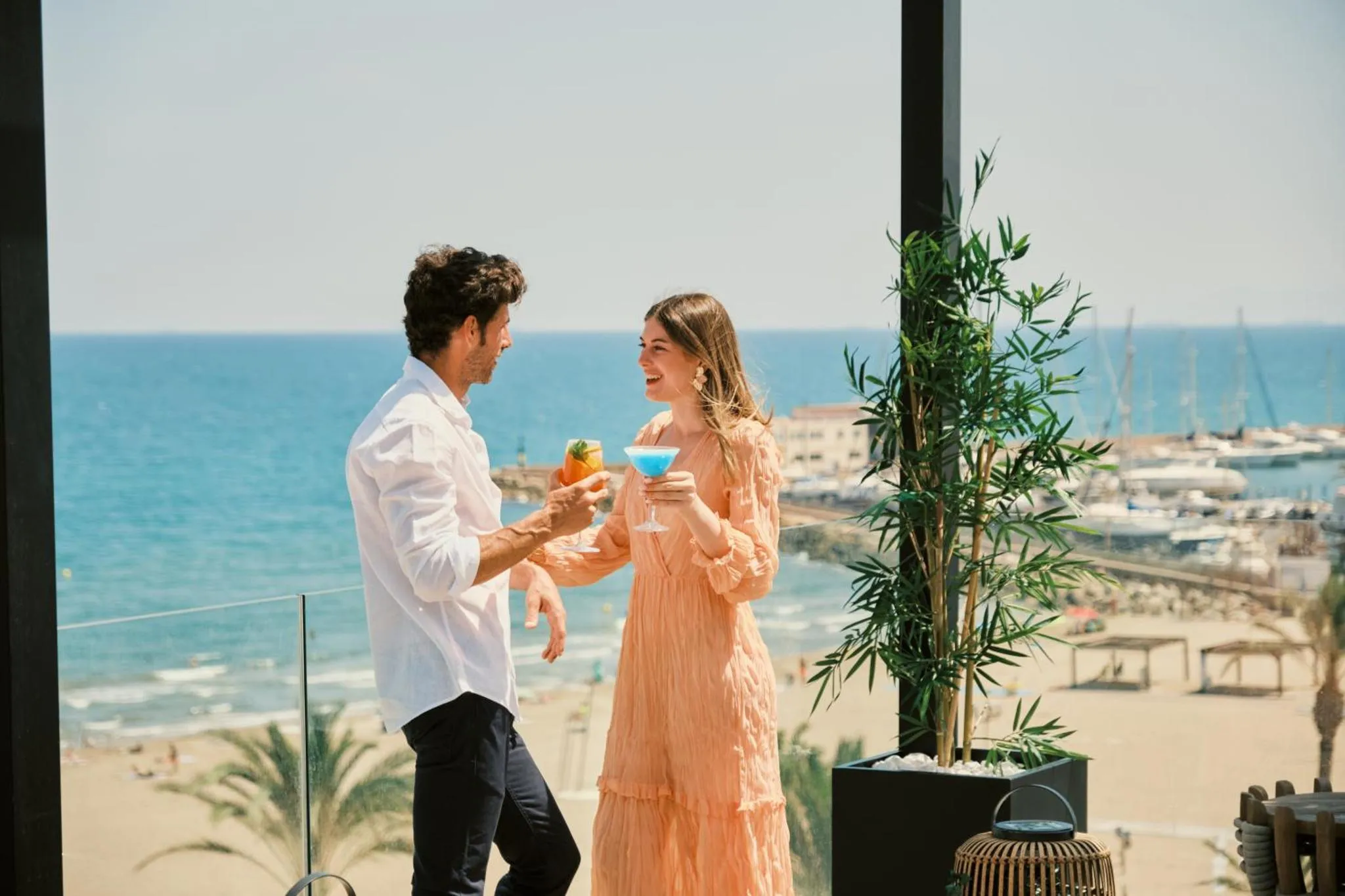 Un couple échange des cocktails sur une terrasse ensoleillée avec vue sur la mer et un port de plaisance.