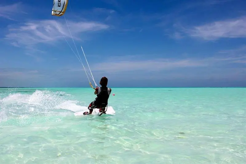 Une personne pratique le kitesurf sur une eau turquoise peu profonde.
