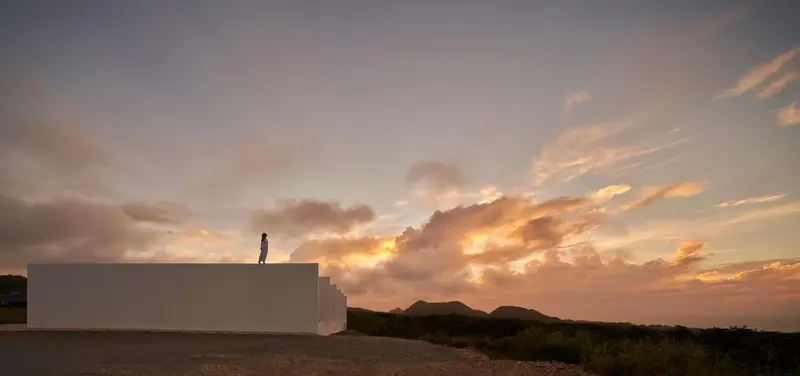 Ciel orangé au coucher du soleil sur une structure blanche et un paysage montagneux.