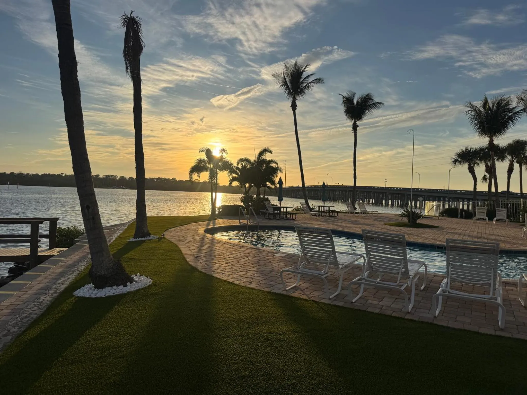 Transats blancs disposés autour d'une piscine au bord de l'eau, avec des palmiers et un ciel orangé au coucher du soleil.