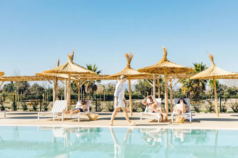 Des transats sont disposés au bord d'une piscine sous des parasols en chaume.
