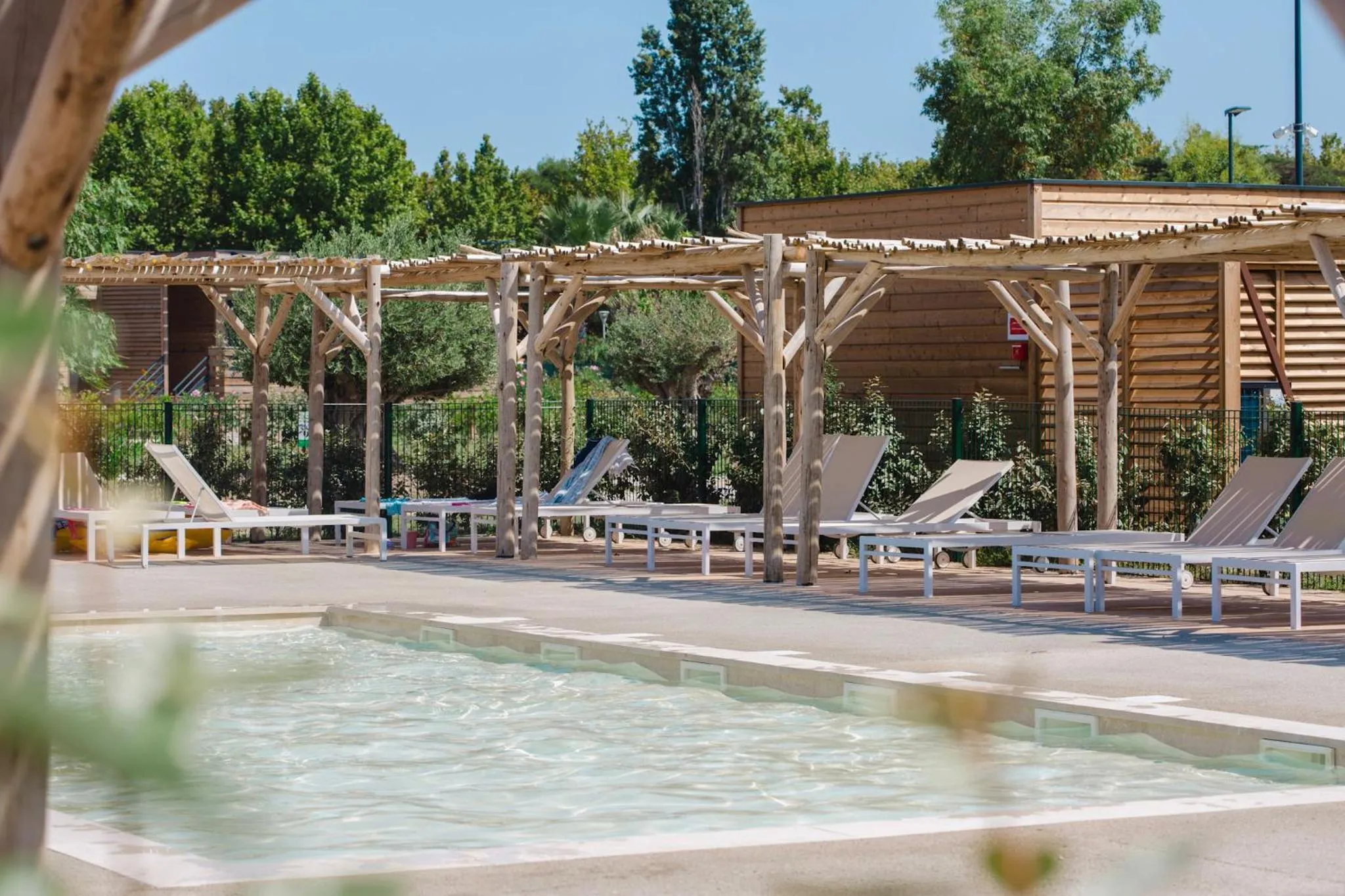 Transats blancs alignés sous une pergola en bois près d'une piscine.