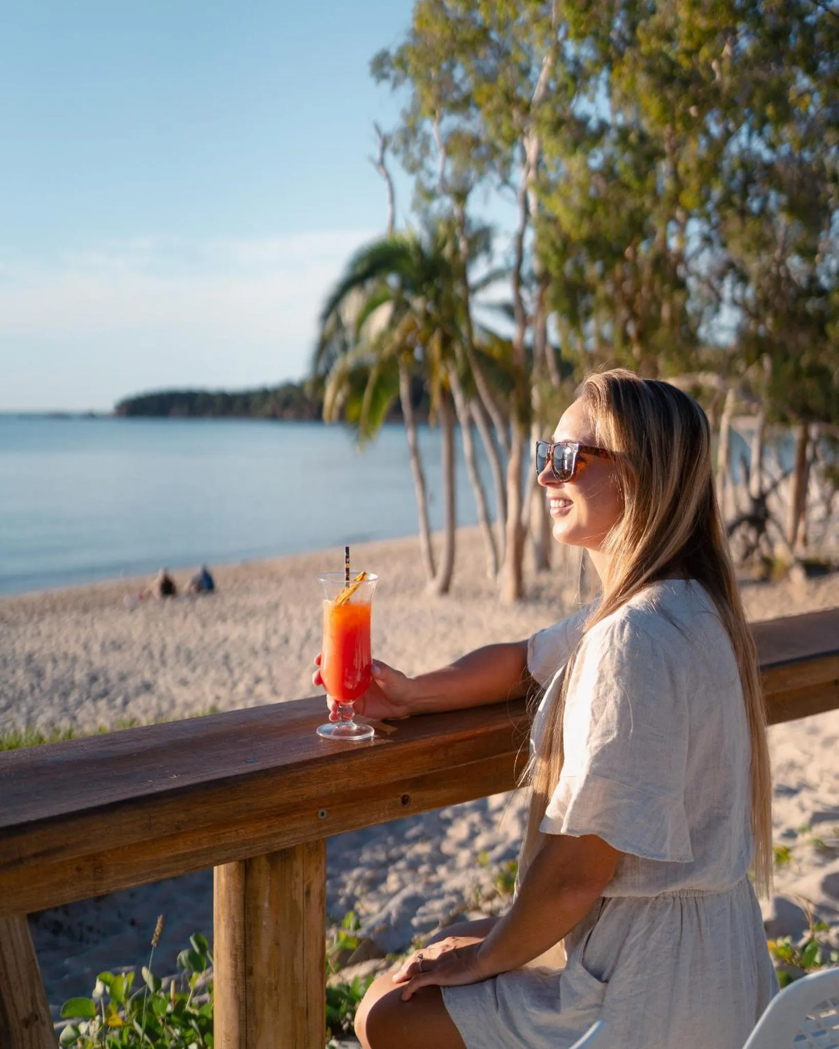 Une femme assise à une table en bois, face à la mer, déguste une boisson colorée.