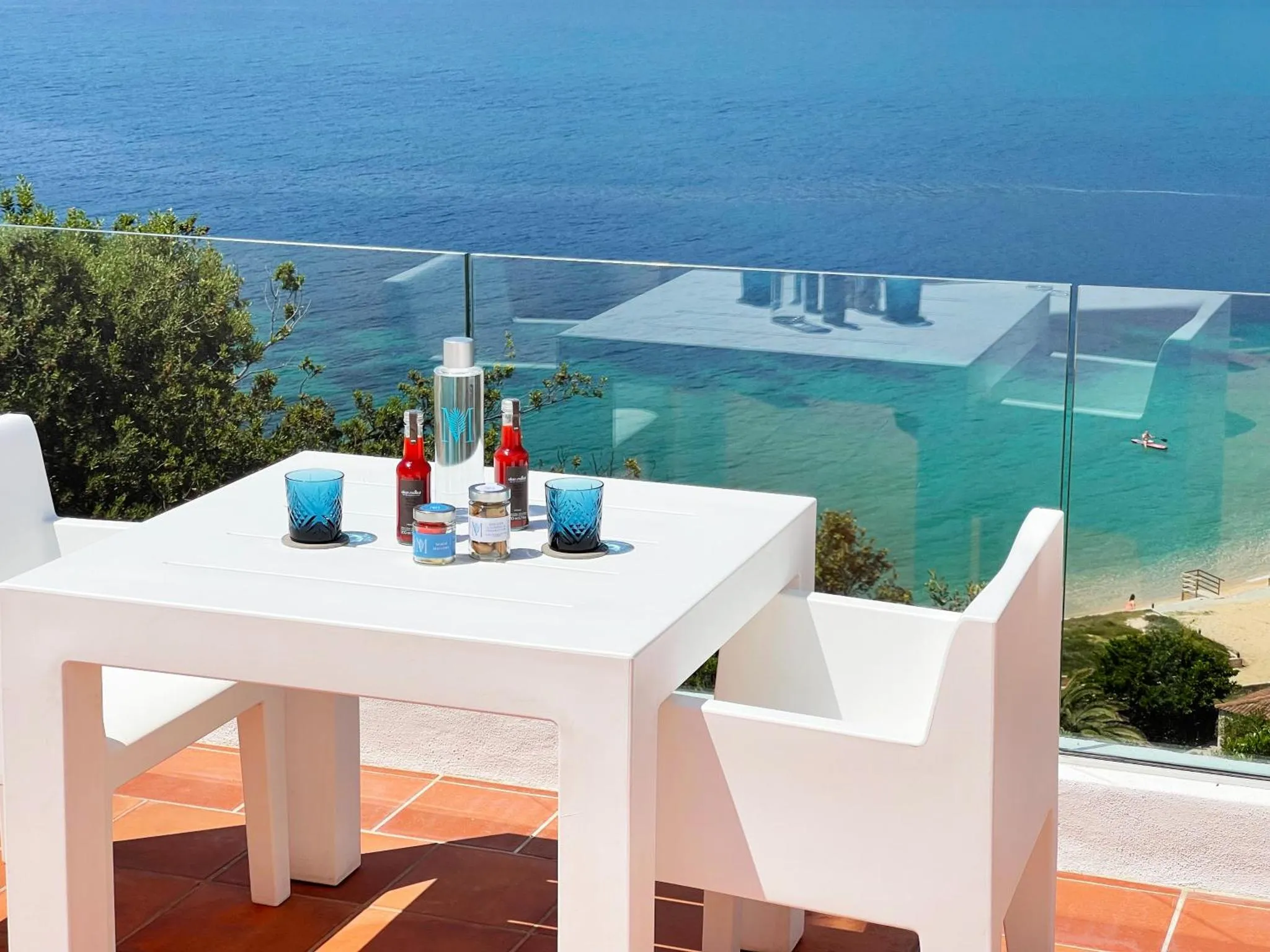 Une table blanche avec des boissons et des collations est mise en scène sur une terrasse ensoleillée avec vue sur la mer.