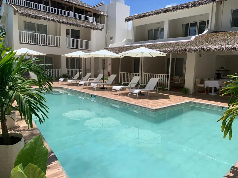 Une piscine turquoise bordée de transats et de parasols devant un bâtiment blanc à l'architecture tropicale.