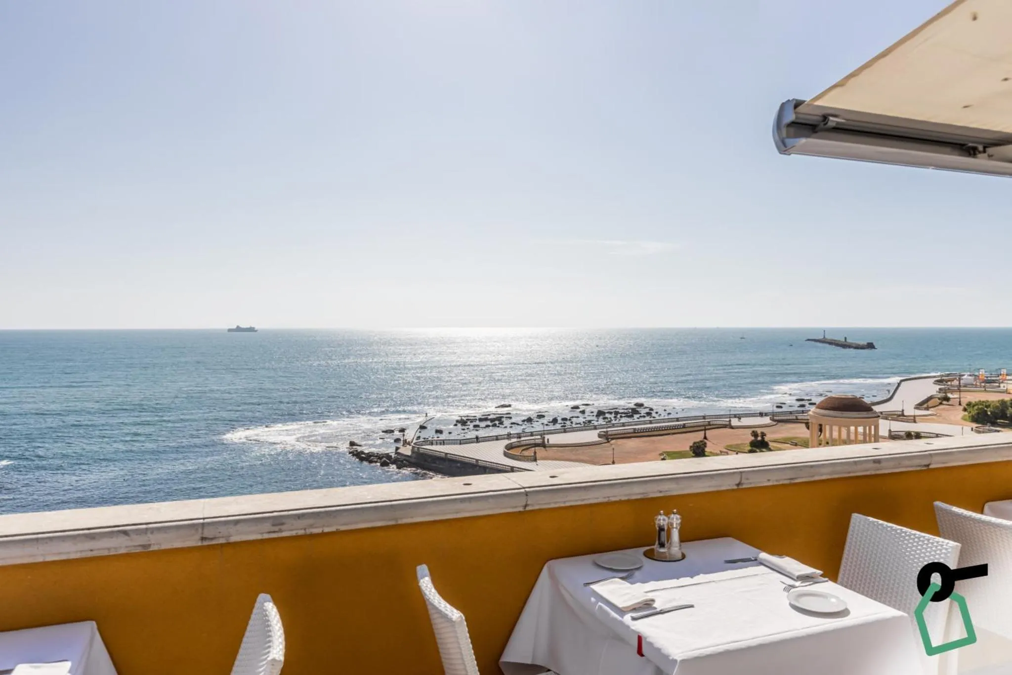 Une terrasse avec des tables dressées offre une vue sur la mer scintillante et la côte rocheuse.
