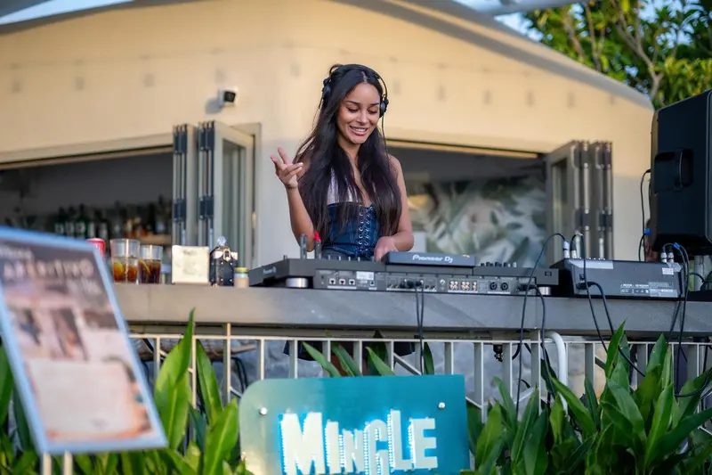 Une jeune femme souriante mixe de la musique devant un bar en extérieur.