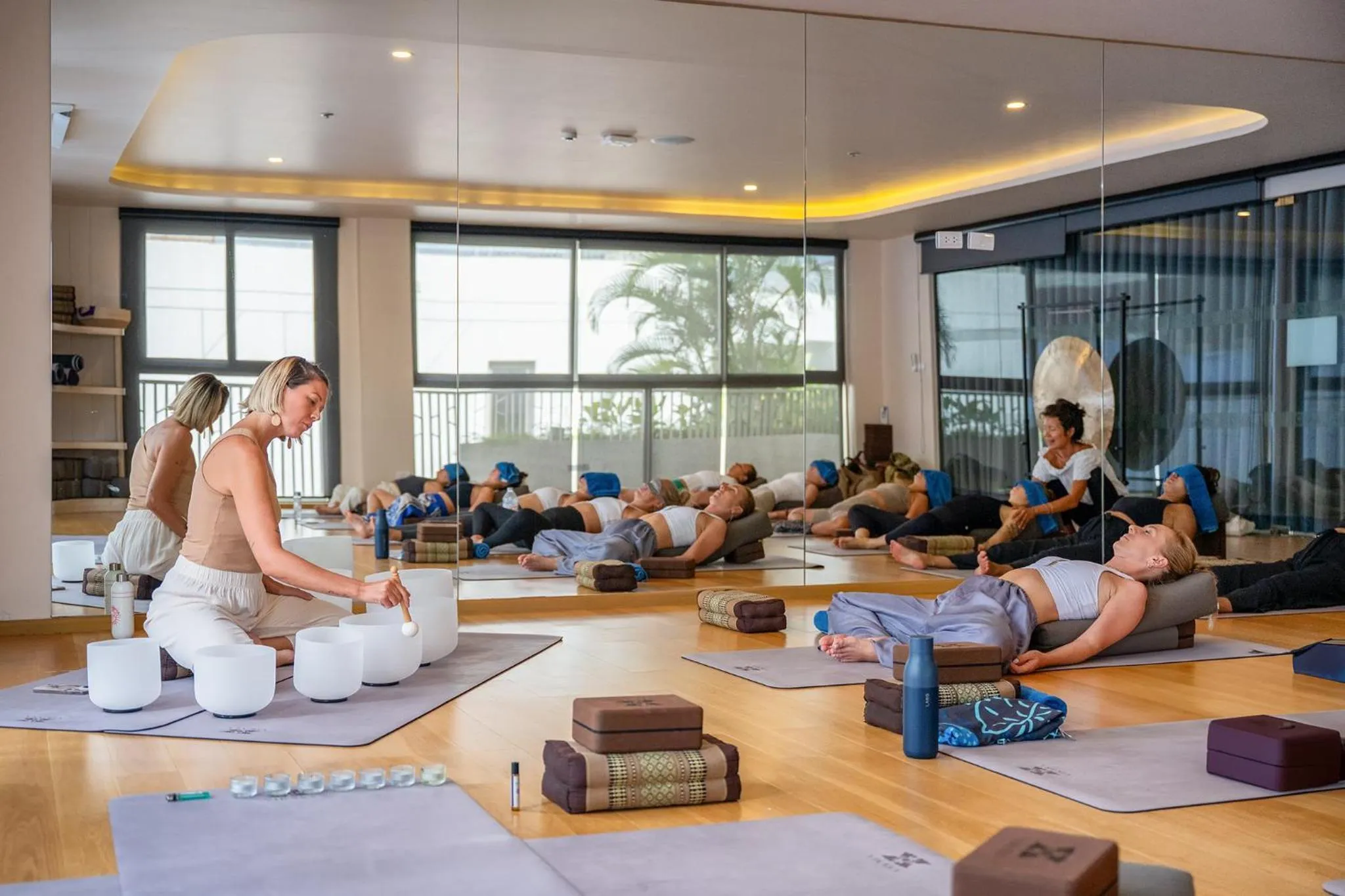 Une instructrice guide un groupe de personnes allongées sur des tapis de yoga dans une pièce lumineuse.